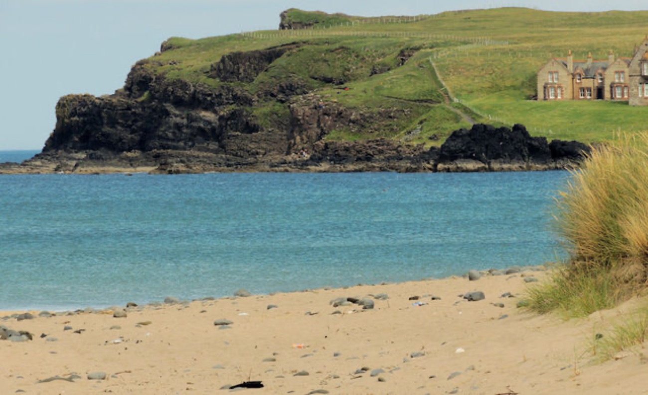 The sandy strand at Portballintrae with grassy dunes and a headland house across the calm bay