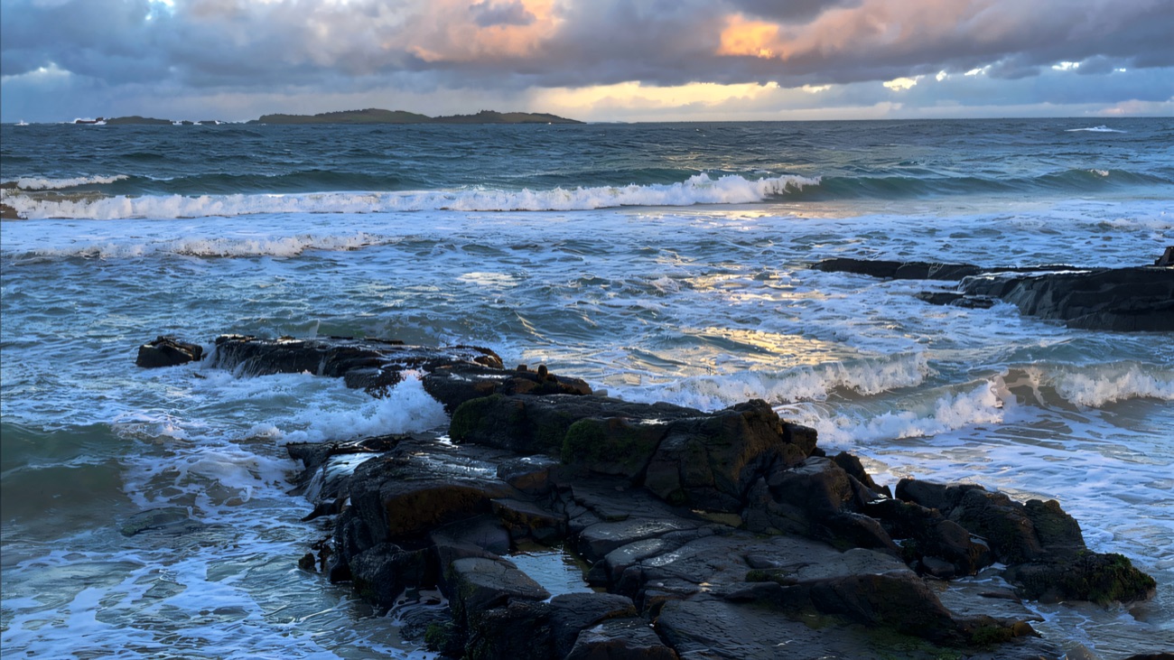 Surf rolling over the dark rock shelves with the Skerries islands low on the horizon.