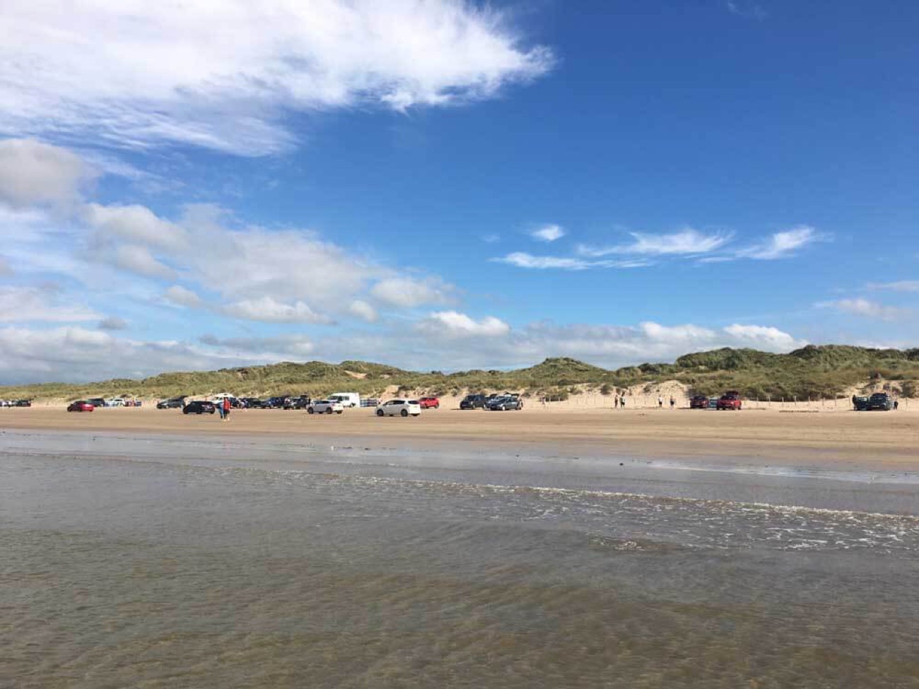 A long line of cars parked on the firm sand at Portstewart Strand under a bright blue sky, with the dunes rising behind and the tide in the foreground