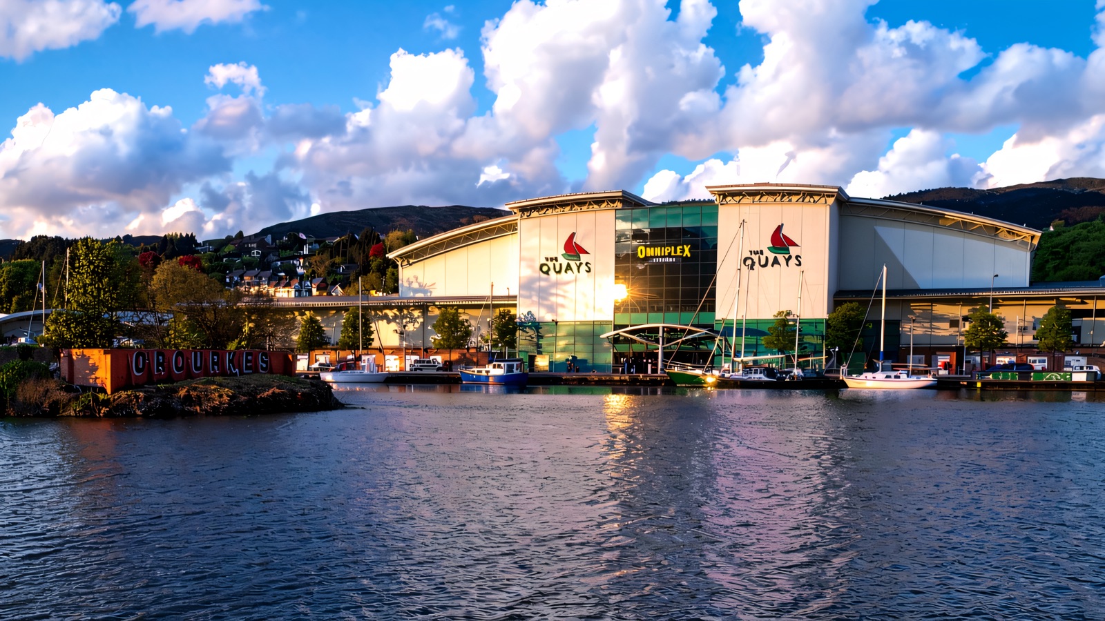The Quays Shopping Centre and its sail logo seen across the water of the Albert Basin, with boats moored in front and the Mourne Mountains behind