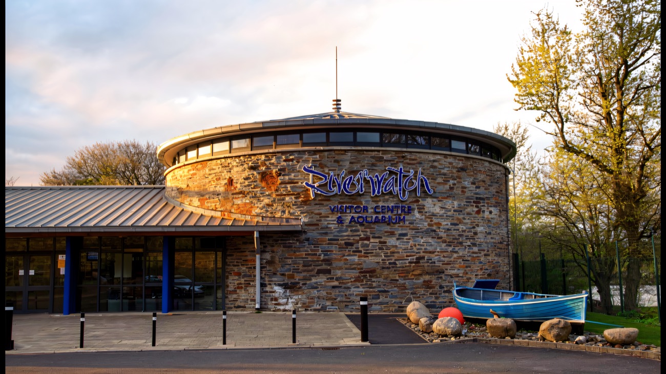 The circular stone Riverwatch visitor centre building from outside, with a little blue boat by the door and the river beyond