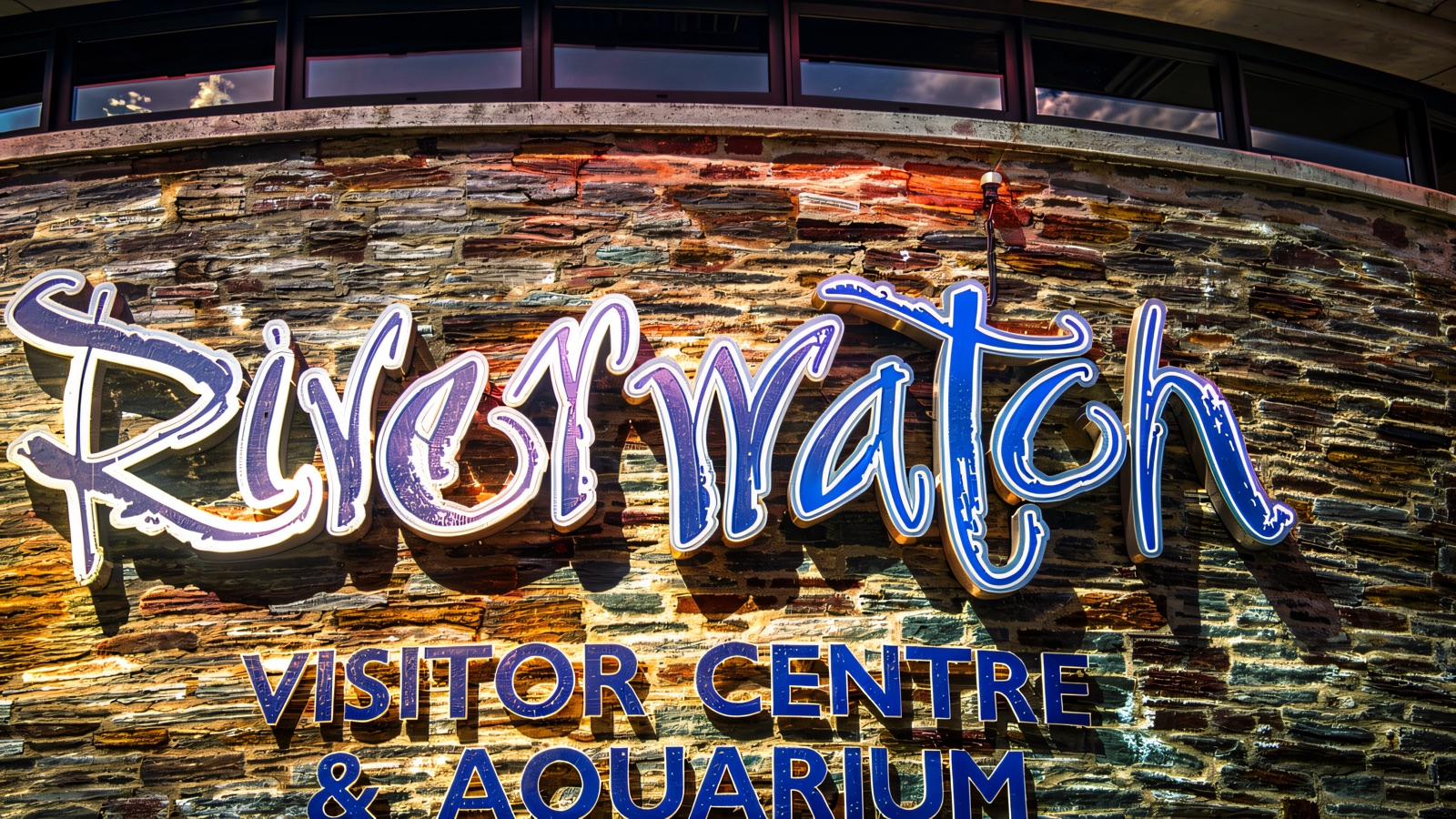 The curved stone frontage of Riverwatch Visitor Centre & Aquarium with its purple-and-blue signage lit up