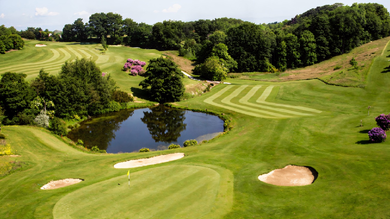 A parkland golf hole with a pond, bunkers and a flag on the green