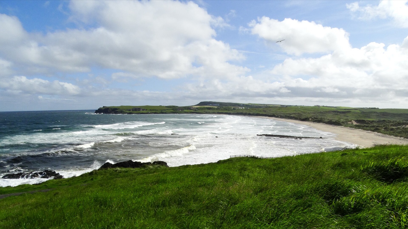 The wide sweep of Runkerry Strand near Bushmills, waves rolling in below dunes and a green headland under a big cloudy sky
