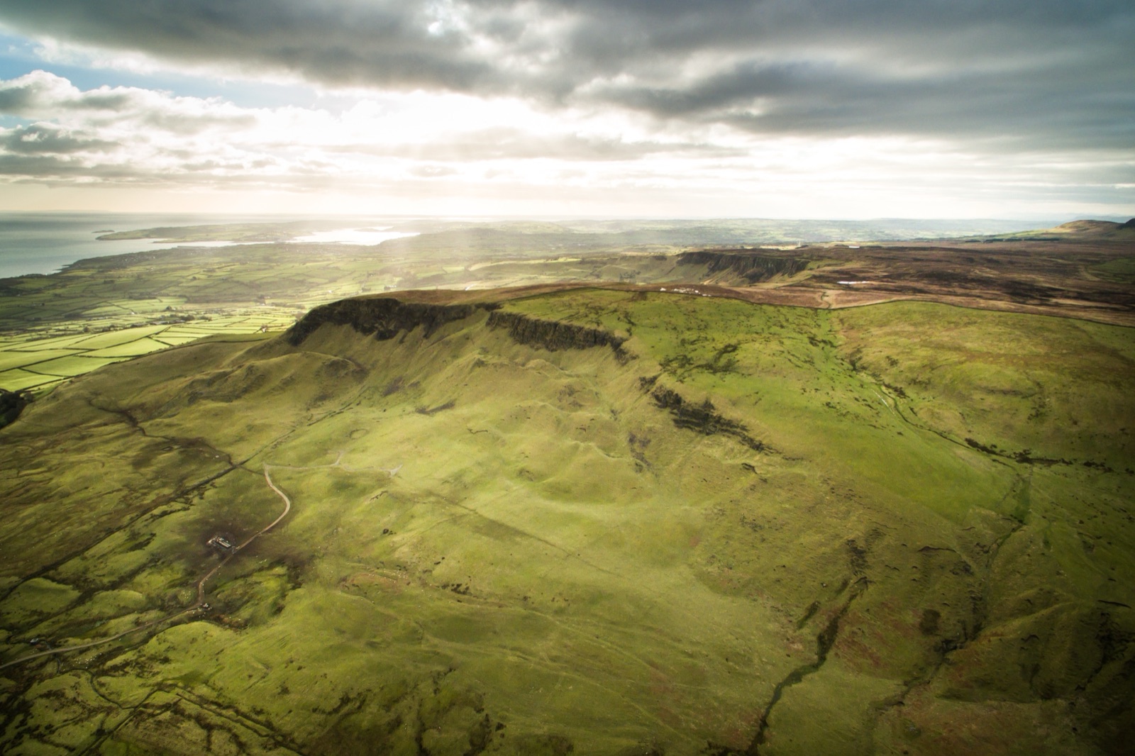 Aerial view of the Sallagh Braes escarpment, a long green cliff line above patchwork fields with the sea beyond