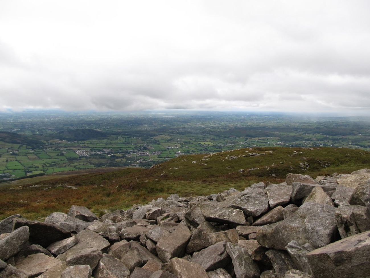 The tumbled stones of the summit cairn with a wide patchwork view over South Armagh below.