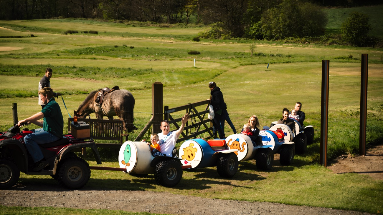 Children riding the colourful barrel-train pulled by a quad, with ponies on the green field behind