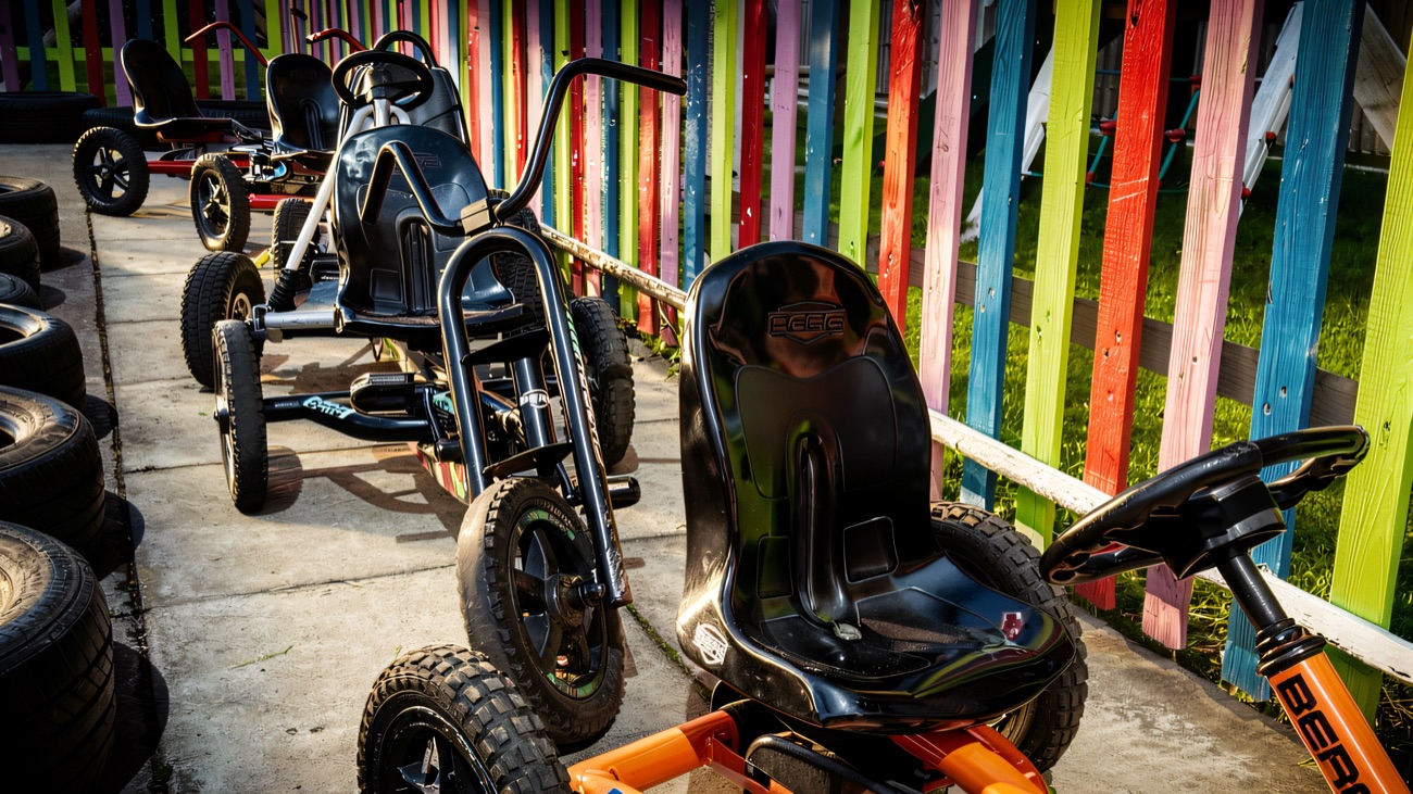 A line of pedal go-karts parked beside a rainbow-painted fence in the sunshine