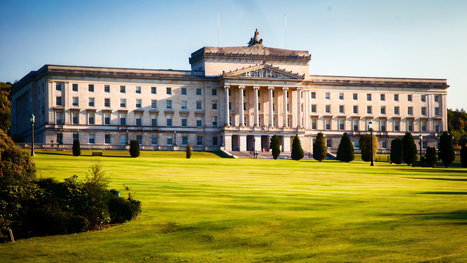The grand white classical Parliament Buildings at Stormont across a wide green lawn under a blue sky