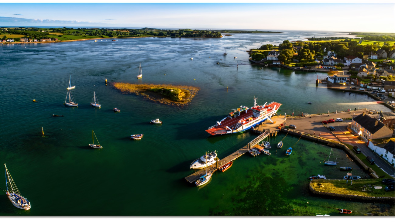 An aerial view of Strangford harbour, the ferry berthed and yachts scattered across the clear water