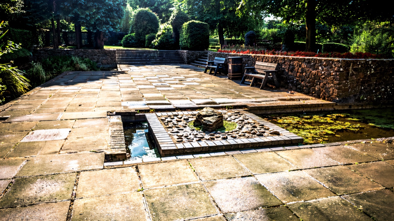 A paved formal garden terrace with a small water feature and beds of red flowers