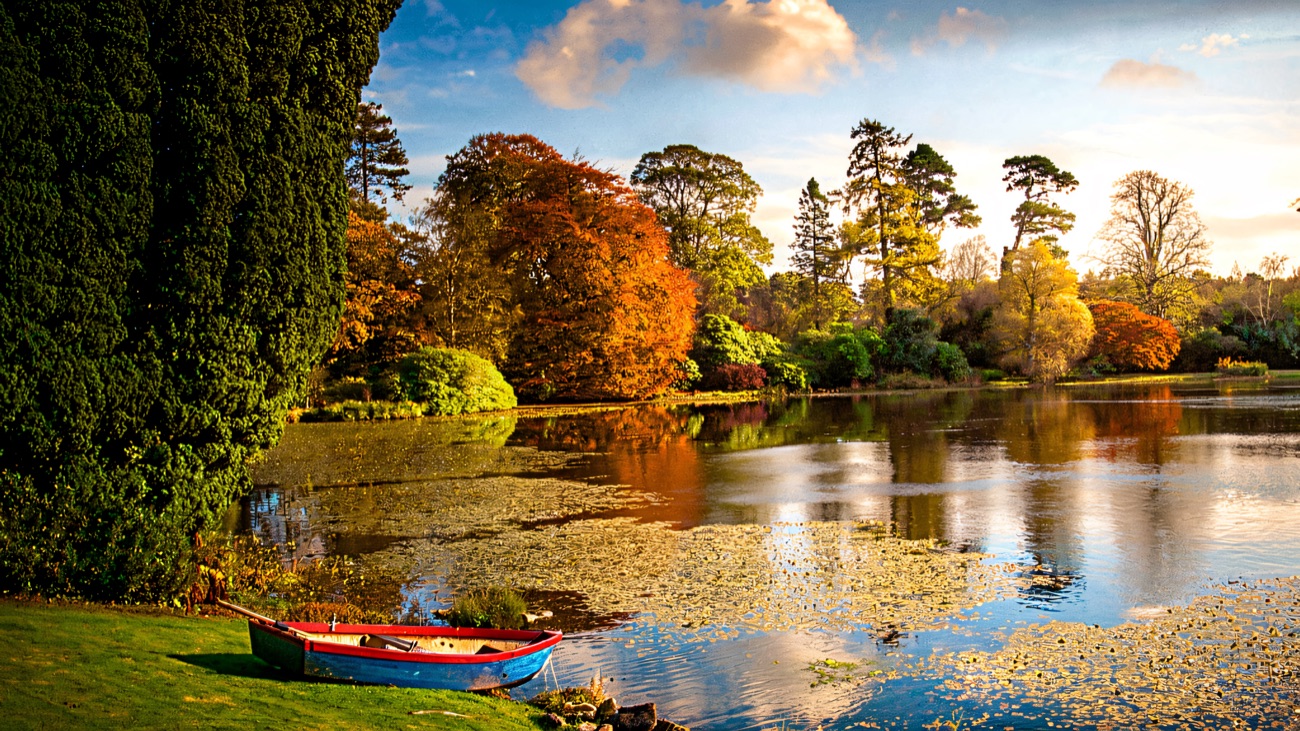 The tree-fringed lough at Tempo Manor with a rowing boat drawn up on the grass and autumn colour reflected in the still water