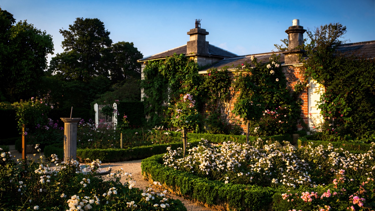 The rose garden at The Argory with a stone sundial, clipped box hedges and beds of pink and white roses under a blue sky