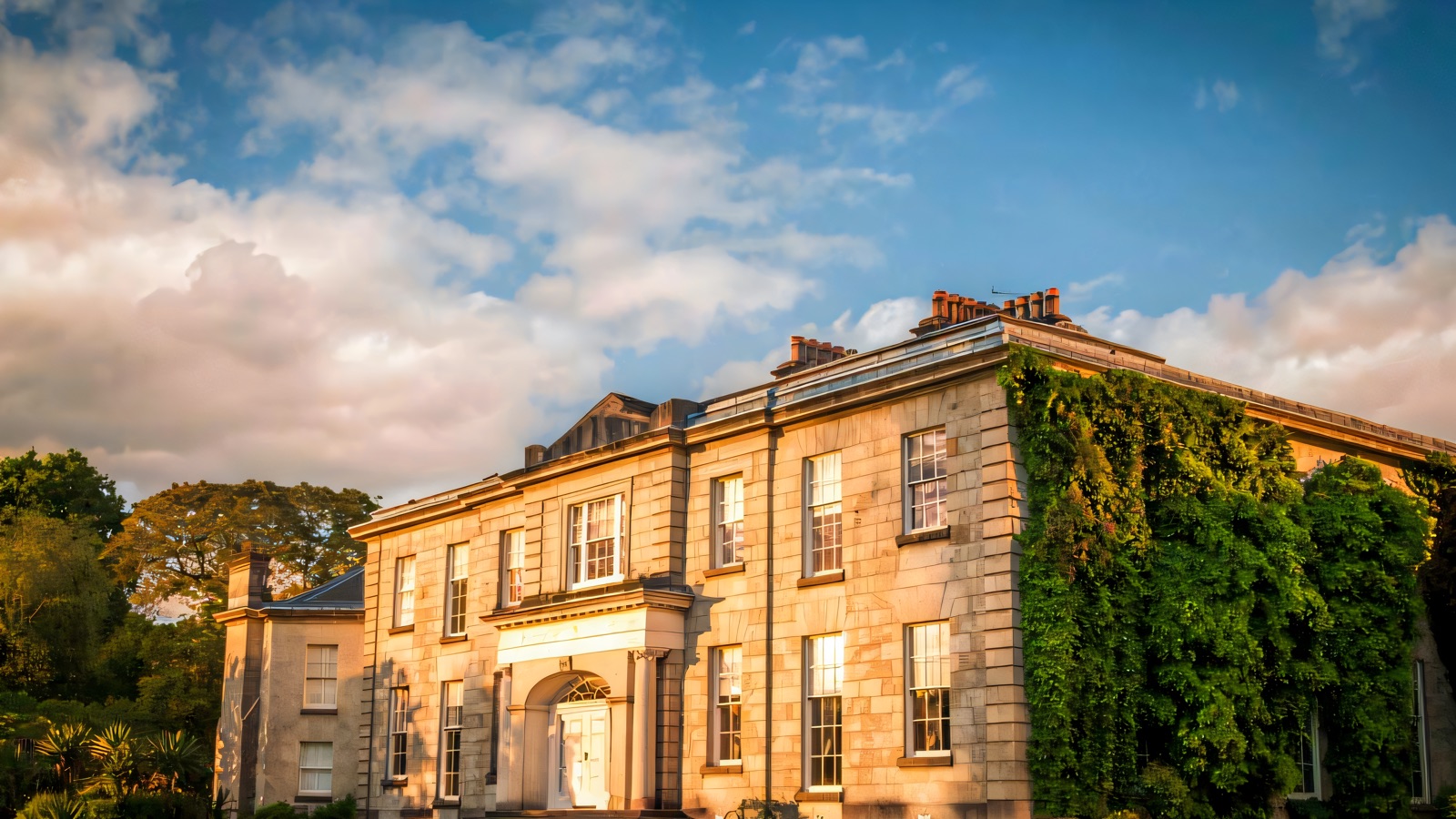 The grey stone Victorian house at The Argory, one wing draped in ivy, seen across a wide green lawn under a blue sky