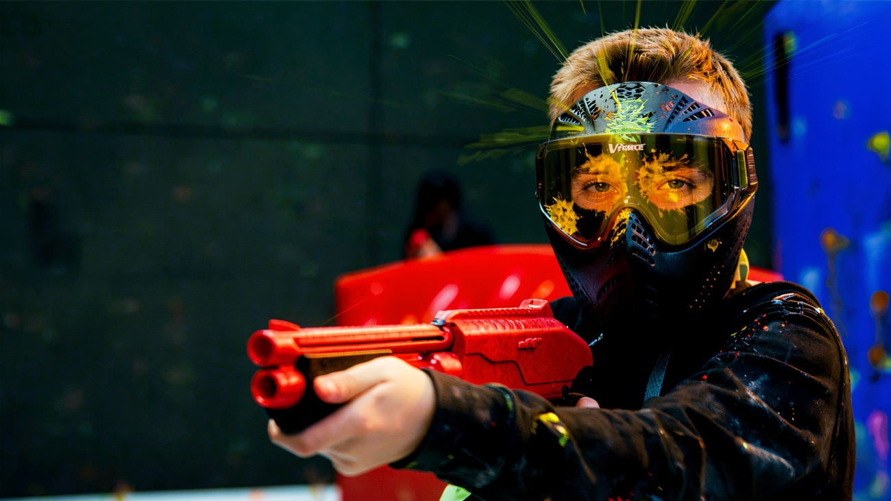 A masked player taking aim, goggles splattered with yellow paint