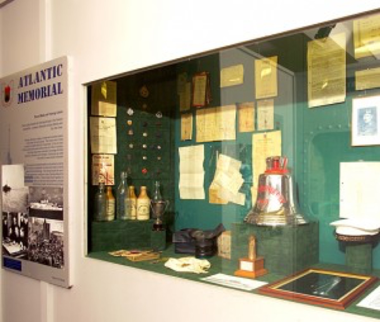 A display case in the Atlantic Memorial exhibition holding a ship's bell, wartime documents and small objects, beside an Atlantic Memorial storyboard
