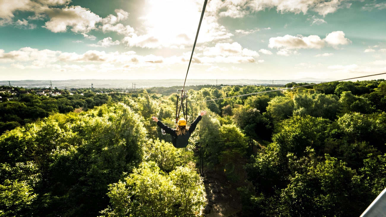 A zip liner flying out over the green treetops of Colin Glen with Belfast in the distance