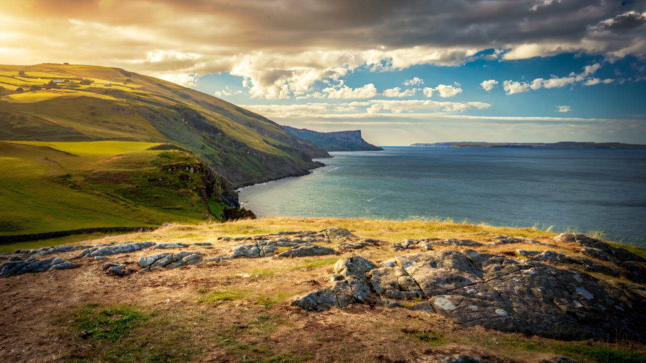 A rocky headland foreground, Fair Head rising across the bay in dappled light