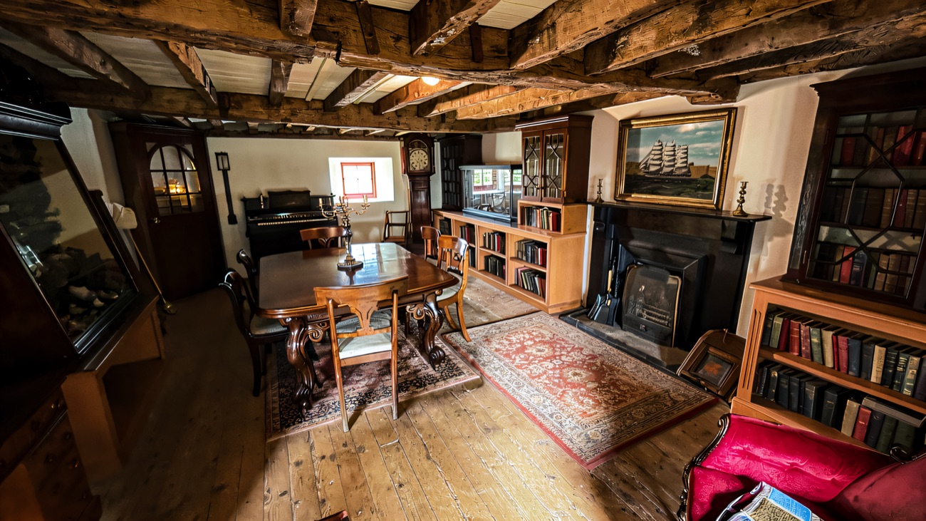 A beamed cottage interior with a cast-iron hearth, table and bookshelves at the Folk Museum