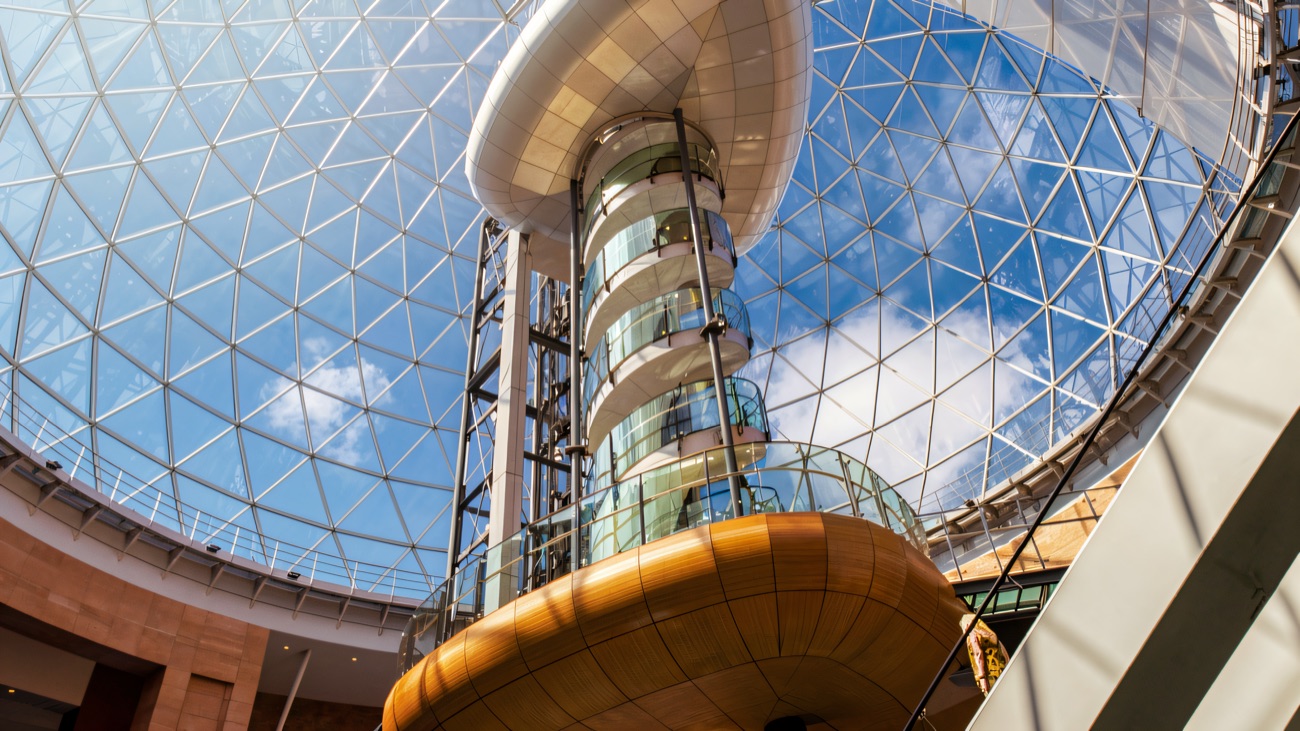 Looking straight up through the atrium of Victoria Square at the glass dome, with the lift tower and viewing pod climbing towards blue sky
