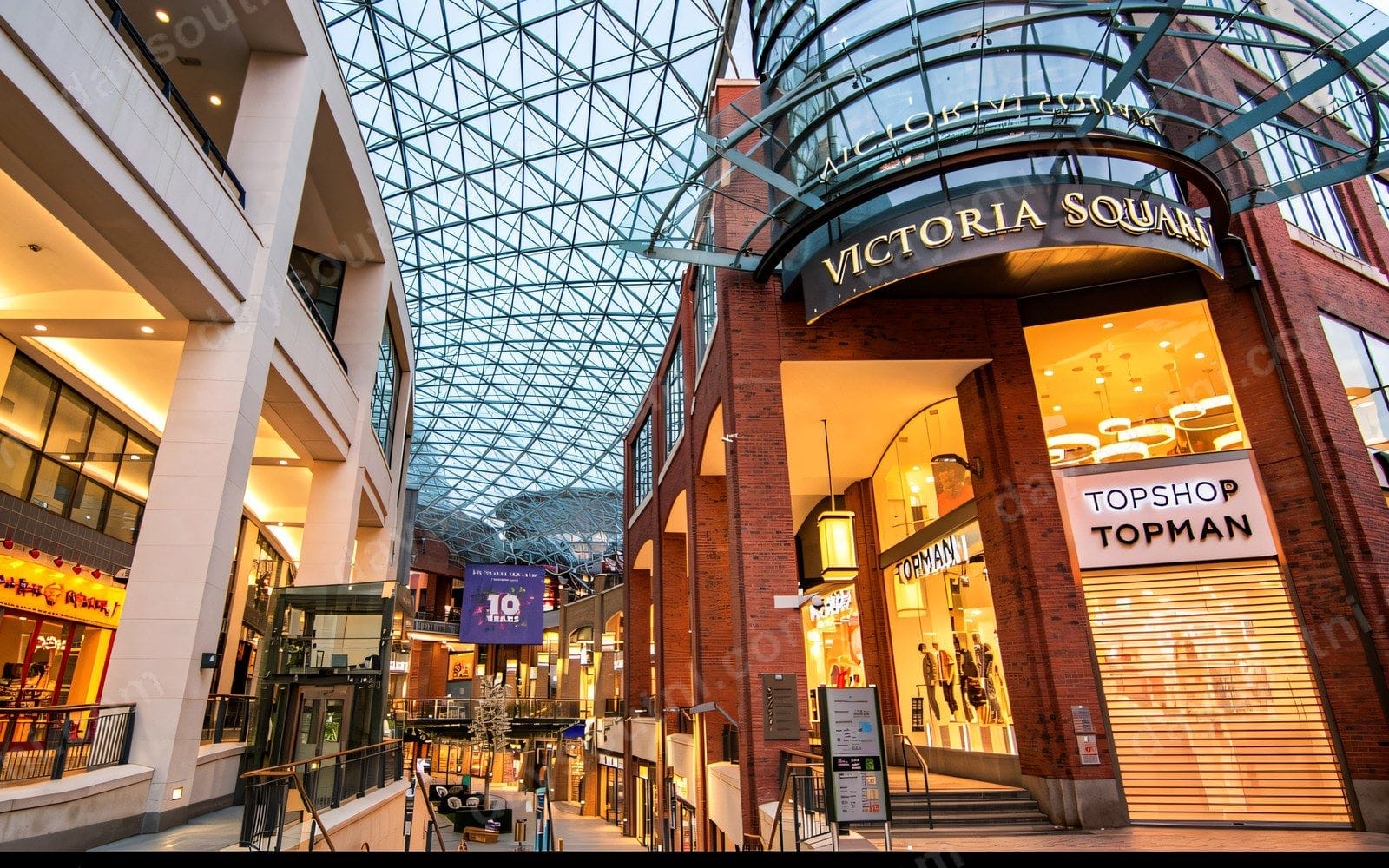 Inside Victoria Square shopping centre, with the steel lattice of the glass roof sweeping over the red-brick shopfronts and upper walkways