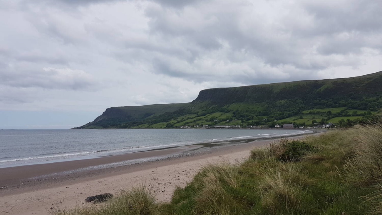 Waterfoot beach curving toward the village at the foot of Glenariff, with the flat-topped glen cliffs sweeping down to the shore