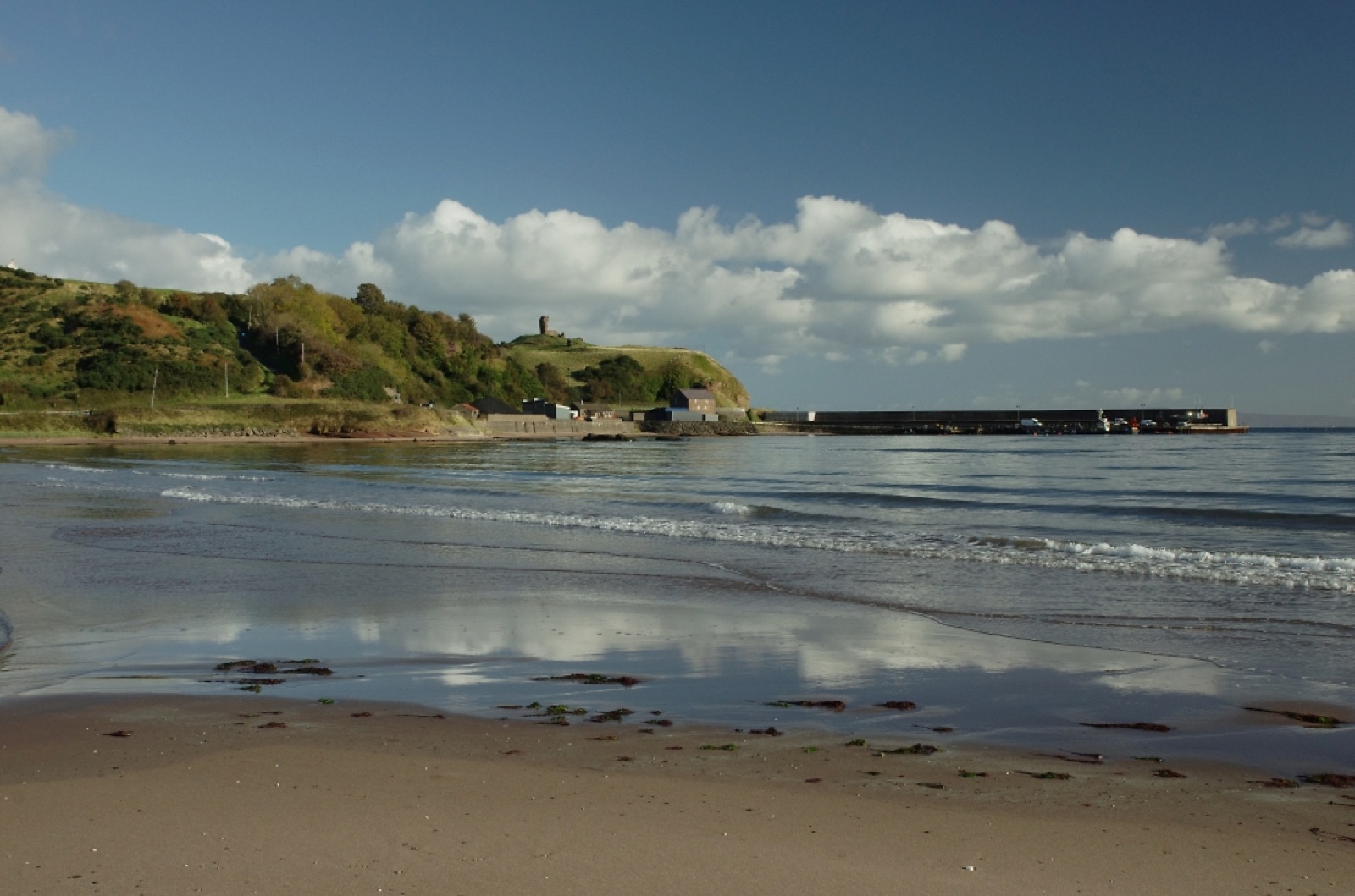 The sandy Red Bay beach at Waterfoot with reflections in the wet sand, the harbour pier and Red Bay Castle on the green headland under a bright sky