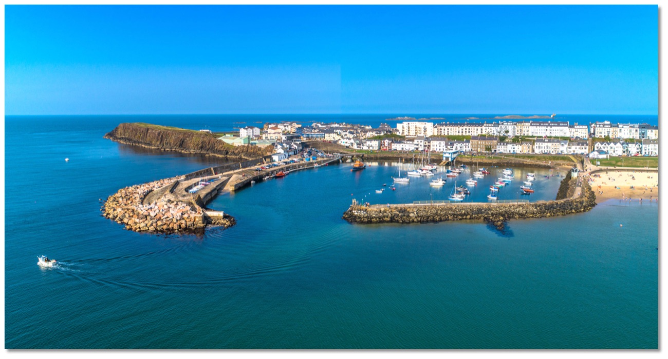 Aerial view of Portrush harbour with small boats moored inside the stone piers, the town and West Strand beyond
