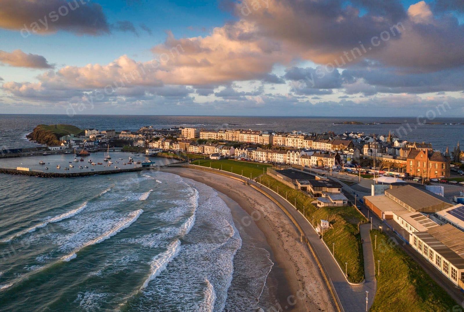 Aerial view of West Strand in Portrush at golden hour, the curve of sandy beach between the harbour and the promenade with the town behind