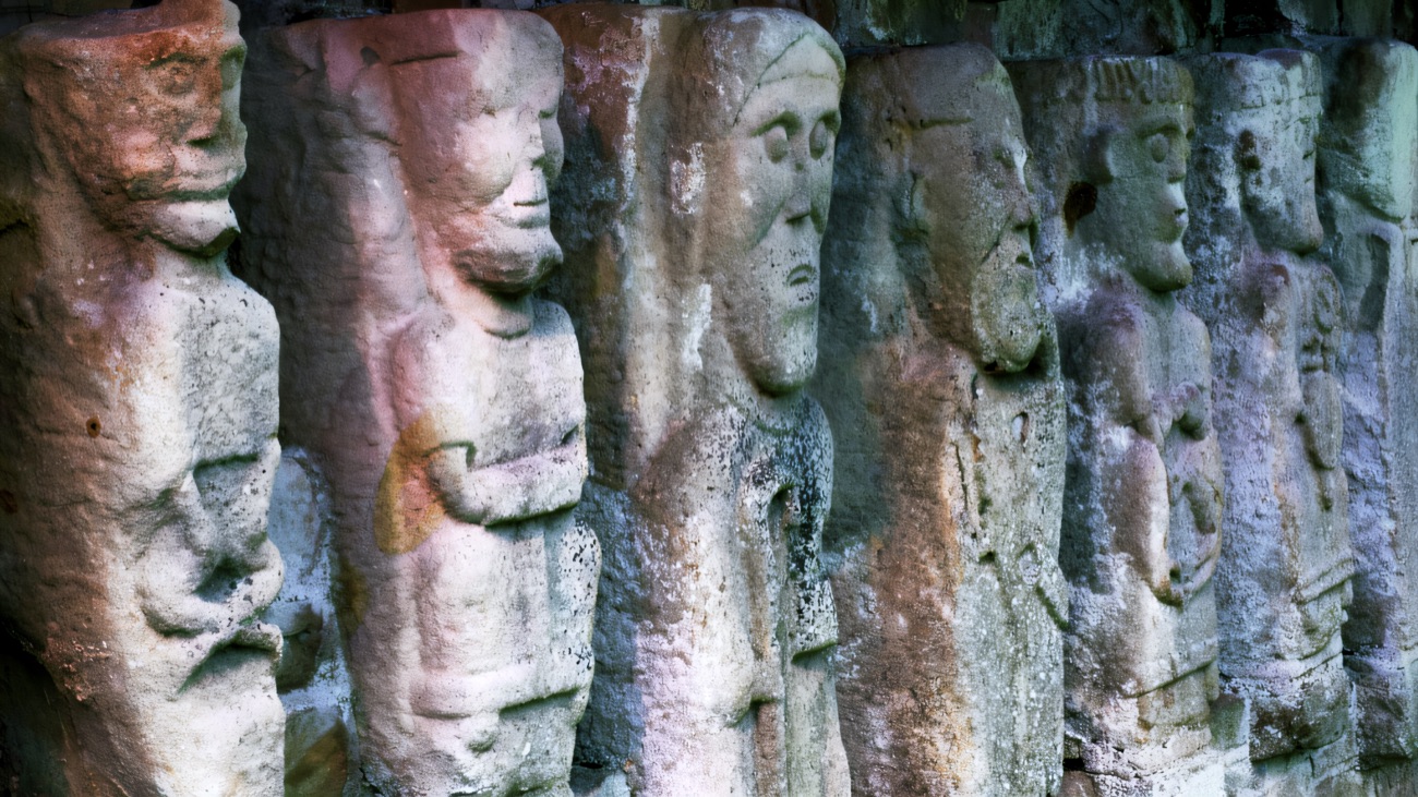 An oblique close-up of the mysterious carved figures, their faces and folded arms in raking light.