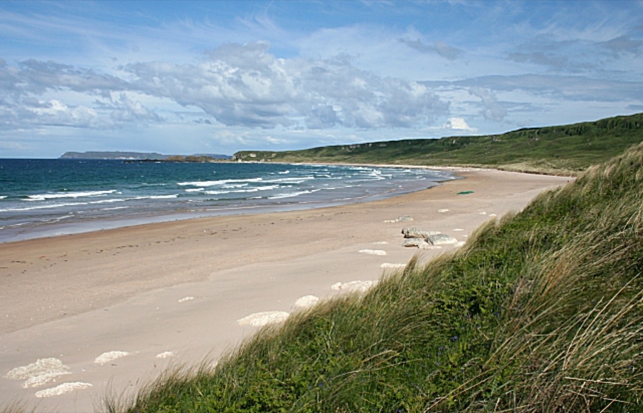 Marram-grass dunes in the foreground looking out along the empty white beach at White Park Bay towards the far headland