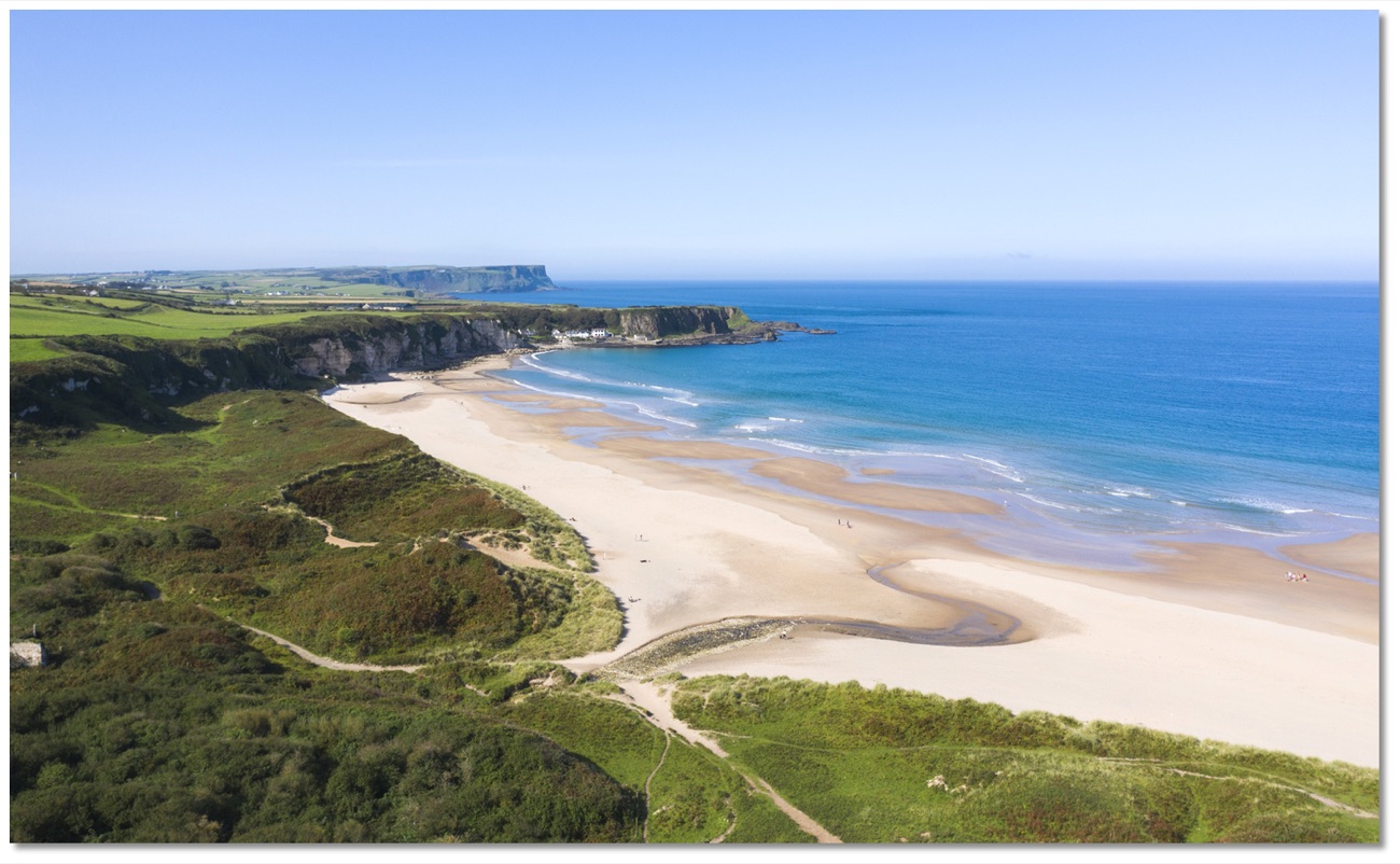 Aerial view of the full curve of White Park Bay at low tide, backed by grassy dunes and white chalk cliffs